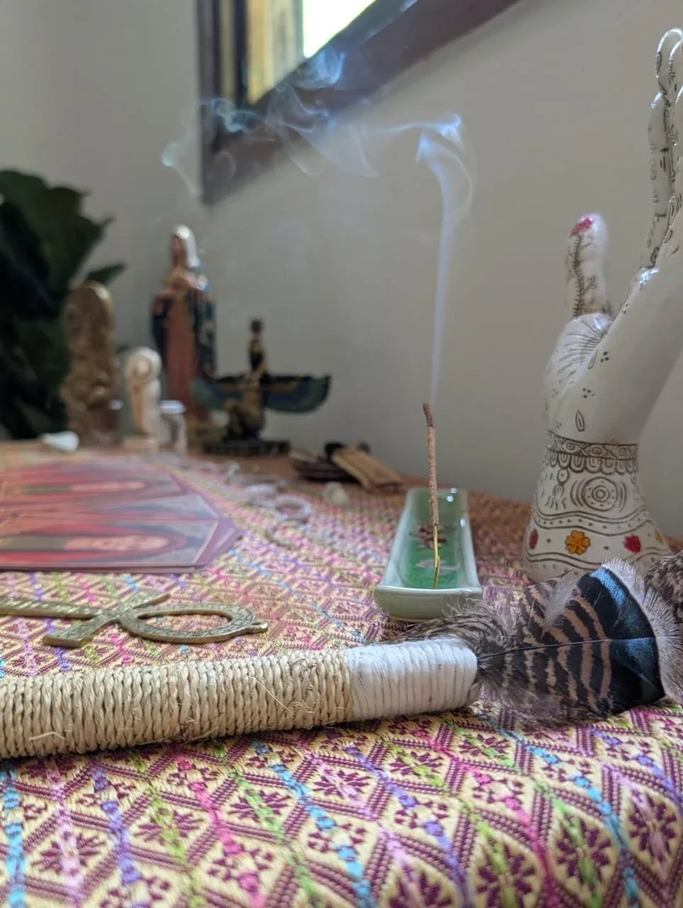 A collection of spiritual and decorative items on a colorful embroidered cloth, including a lit incense stick in a green dish with smoke rising, a white ceramic hand sculpture with painted designs, small figurines, a feather, and some tarot cards.