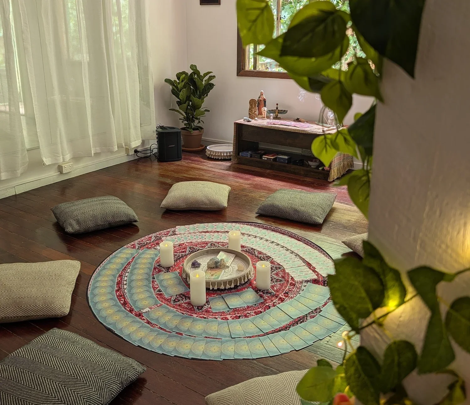 A cozy room with a round mandala-patterned carpet on dark wooden floor, surrounded by floor cushions. On the carpet, there are four candles, crystals, and tarot cards. Against the white wall, a small altar with religious figurines and items is visible, with potted plants nearby. Soft curtains cover a window with sunlight streaming in.