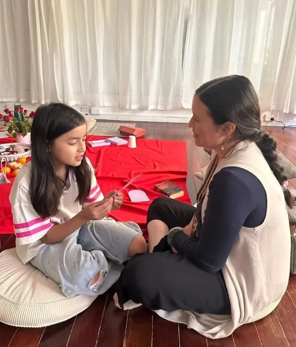 A young girl and an adult woman sitting on the floor facing each other. The girl is holding a card or piece of paper, and the woman is smiling. There are red decorations and flowers on a table behind them, and various items including books, plants, and small objects on the floor near them.