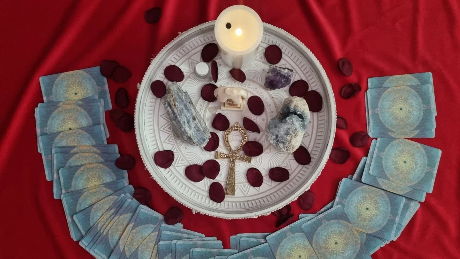 A circular arrangement of tarot or oracle cards with a red backdrop. In the center, there are crystals, a white candle, a small skull, a key pendant, and rose petals scattered around.