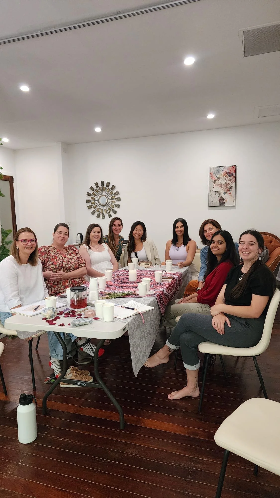 Group of women sitting around a table at a social gathering, decorated with a red and pink tablecloth, candles, and rose petals, in a well-lit room with white walls and wall art.