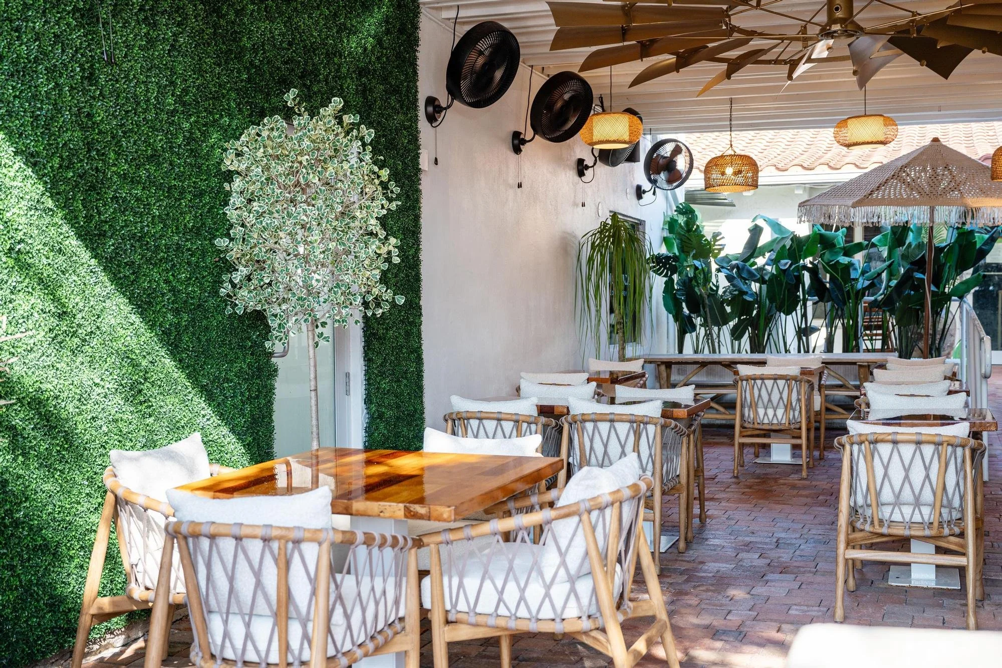Outdoor patio with wooden tables and chairs, lush green plants, and a vertical green hedge wall. Ceiling fans and woven pendant lights hanging from the ceiling, with a large window at the back letting in natural light.