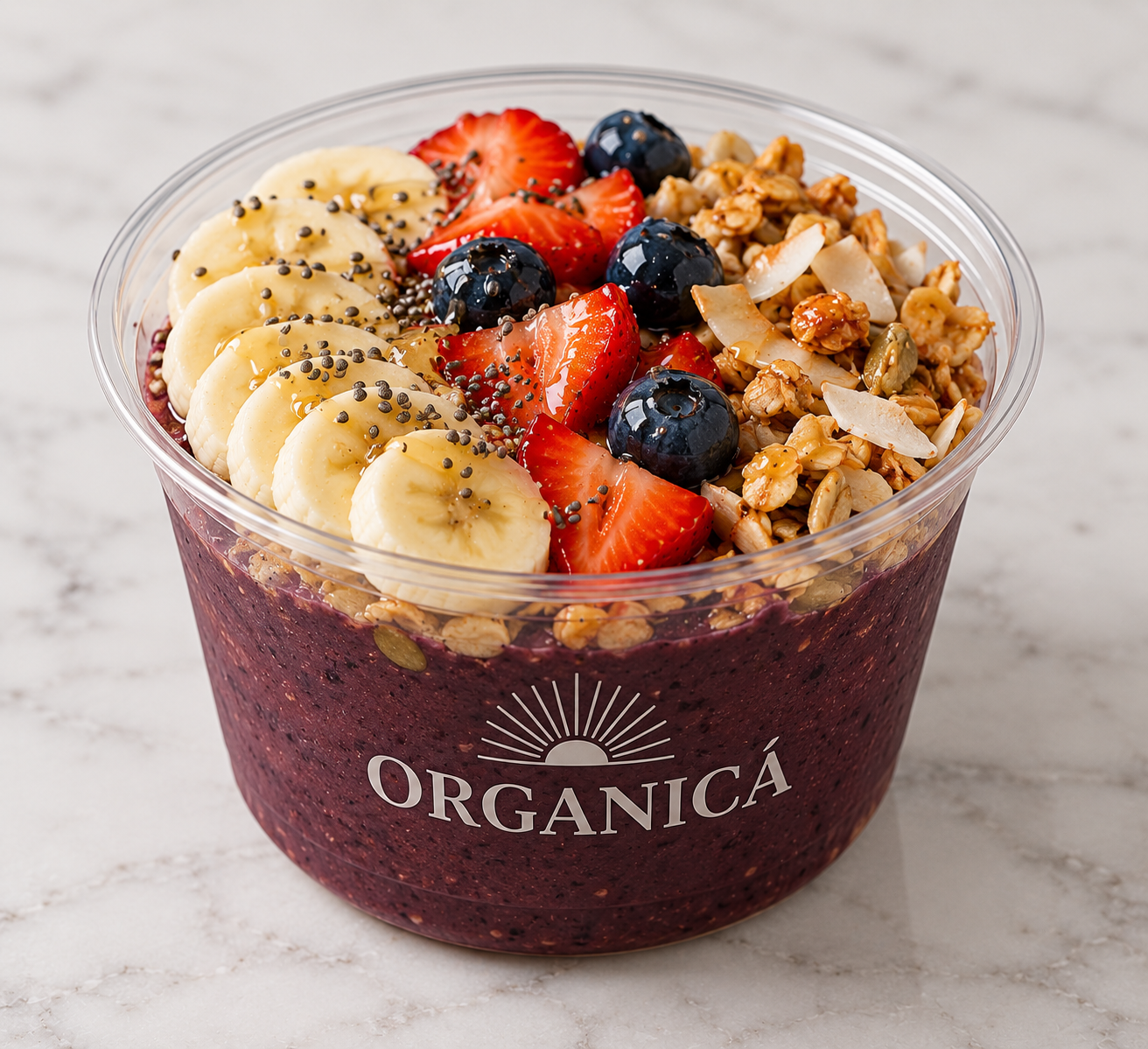 A clear plastic bowl containing a smoothie bowl topped with sliced bananas, strawberries, blueberries, granola, and chia seeds, with the word 'ORGANICÁ' printed on the front of the bowl.