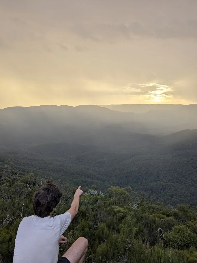 Person sitting on a hillside, pointing toward a sunset or sunrise over a mountain landscape with dense green trees.