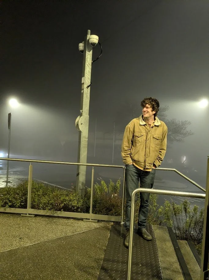 Young man standing outside on a foggy night near a metal railing, with a surveillance camera on a tall pole behind him.