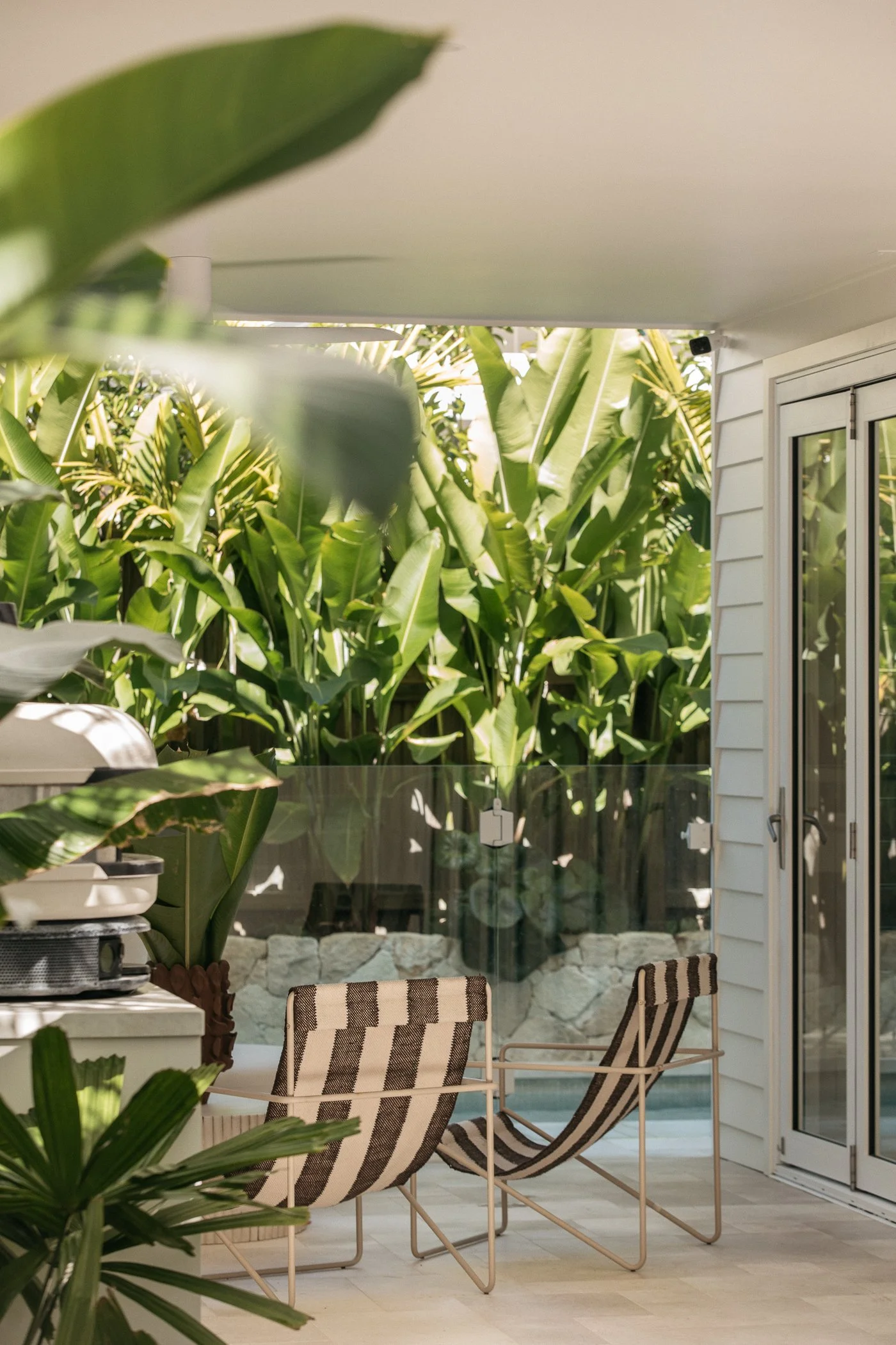 A backyard patio with two striped lounge chairs, large green plants, and a sliding glass door leading into a house.
