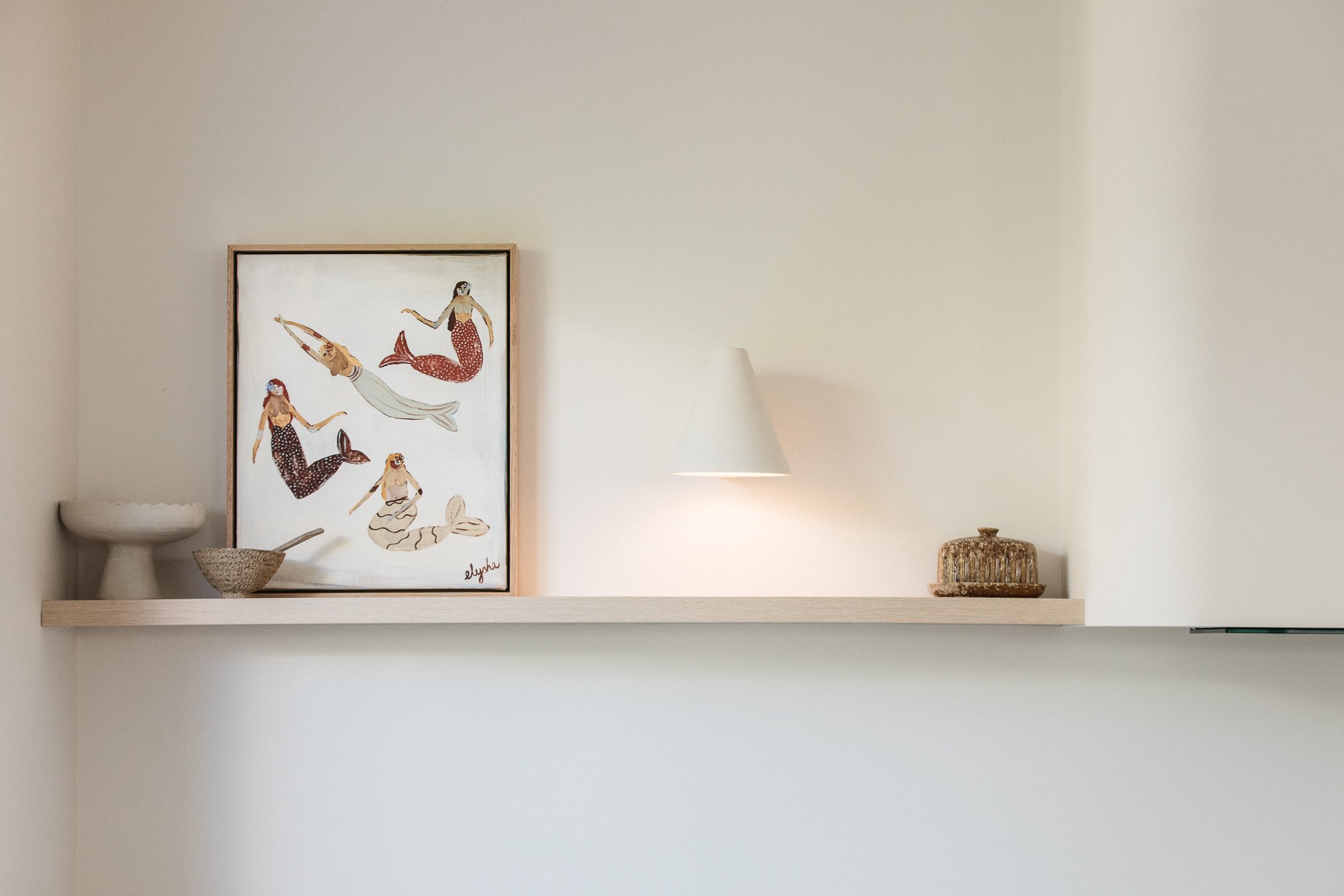 A minimalist interior with a white floating shelf holding a framed illustration of mermaids, a white ceramic bowl, a woven basket, and a wicker container; a white wall and a small lamp are also visible.