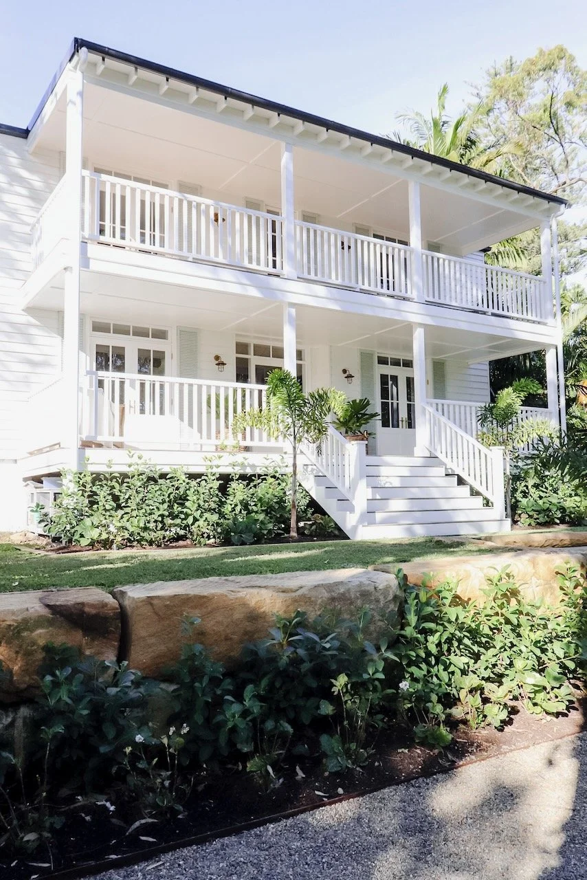 A white two-story house with a large front porch and railing, surrounded by landscaped plants and trees, with stairs leading up to the front door.
