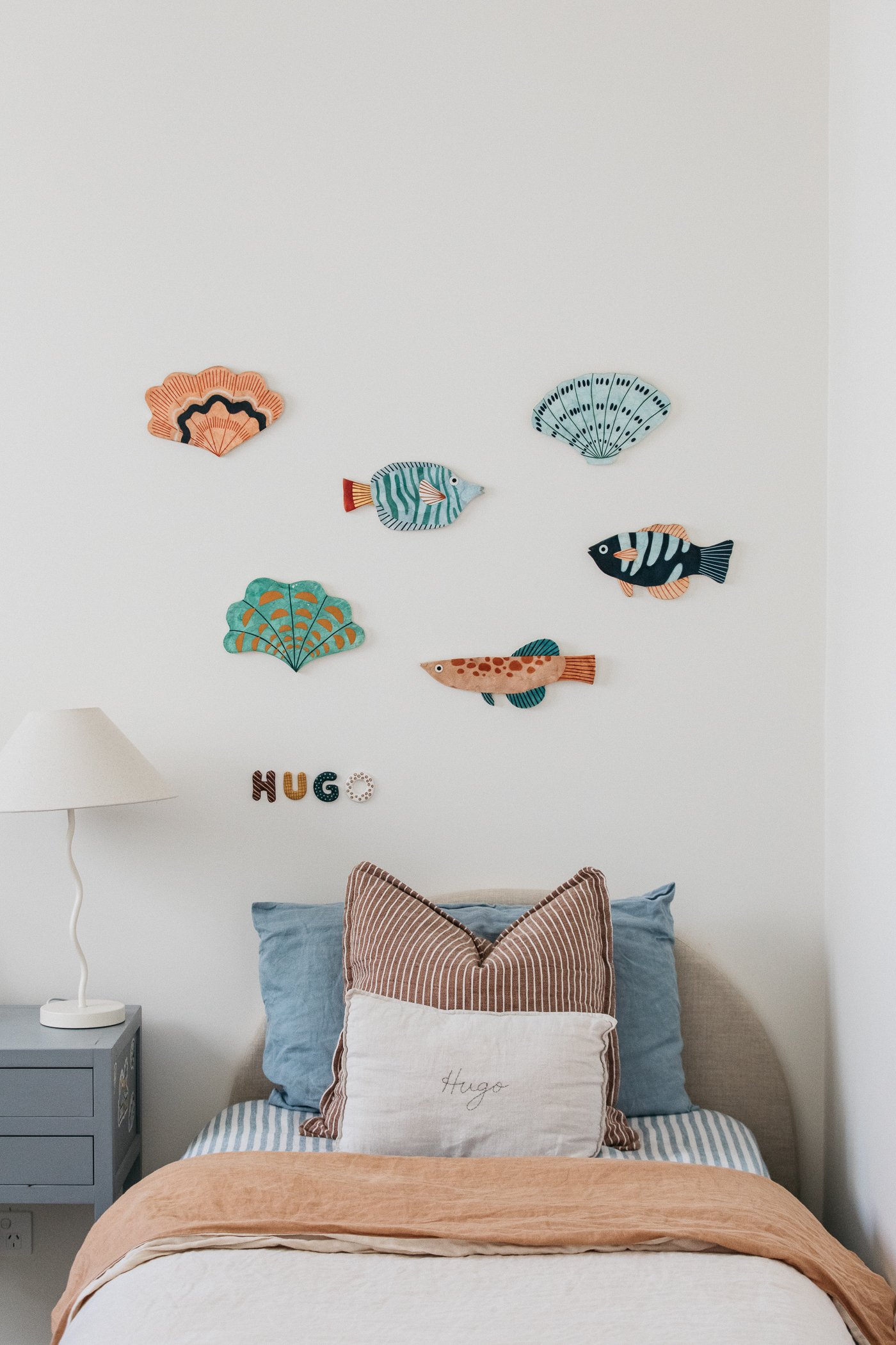 Decorative wall with colorful fish and seashell designs above a bed with pillows, including a personalized pillow with the name "Hugo" embroidered on it.