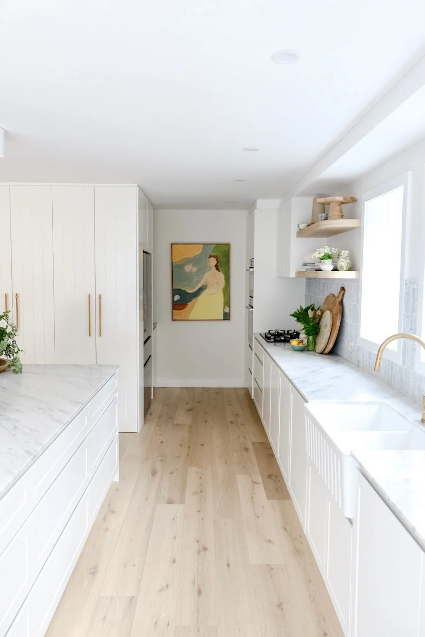 Bright, white kitchen with marble countertops, light wood flooring, and built-in cabinetry, featuring a painting on the wall, open floating shelves, a window, and decorative plants.