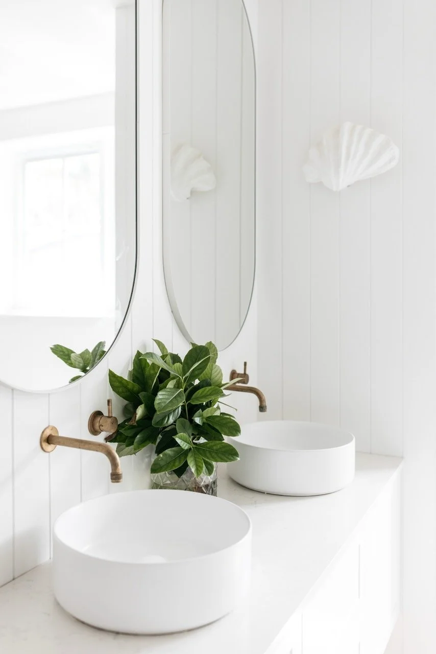 White bathroom vanity with two vessel sinks, a green potted plant, and two mirrors on a white-paneled wall.