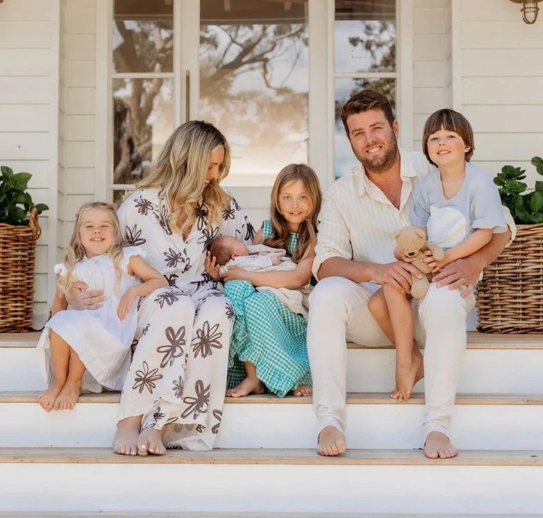 A family sitting on front porch steps, celebrating a newborn with children and parents smiling, holding gifts and stuffed animals, in front of a white house.