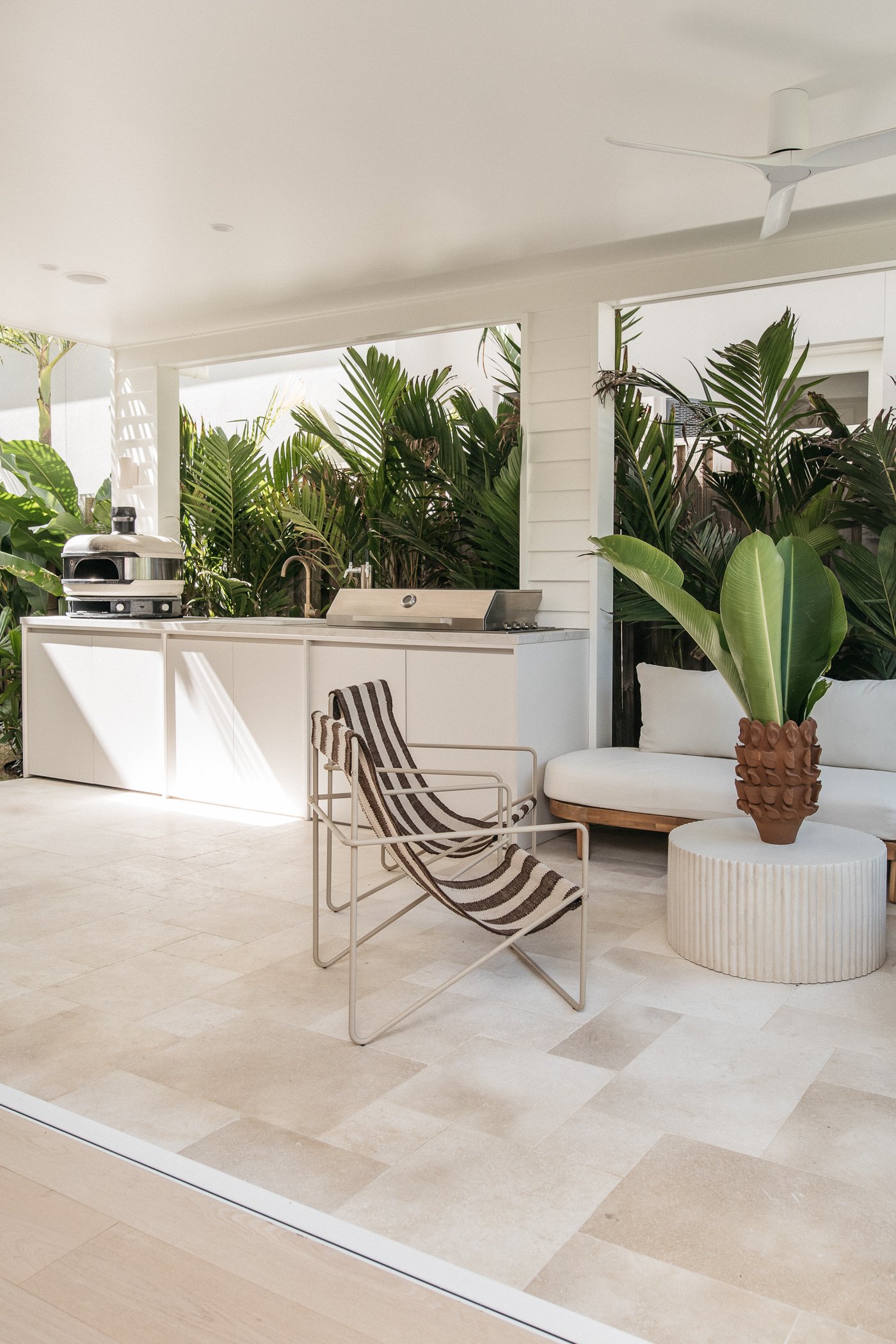 Modern outdoor patio with beige tile flooring, white walls, lush green tropical plants, black and white striped lounge chair, beige cushioned seating, decorative pottery, large window, ceiling fan, and outdoor kitchen area with grill.