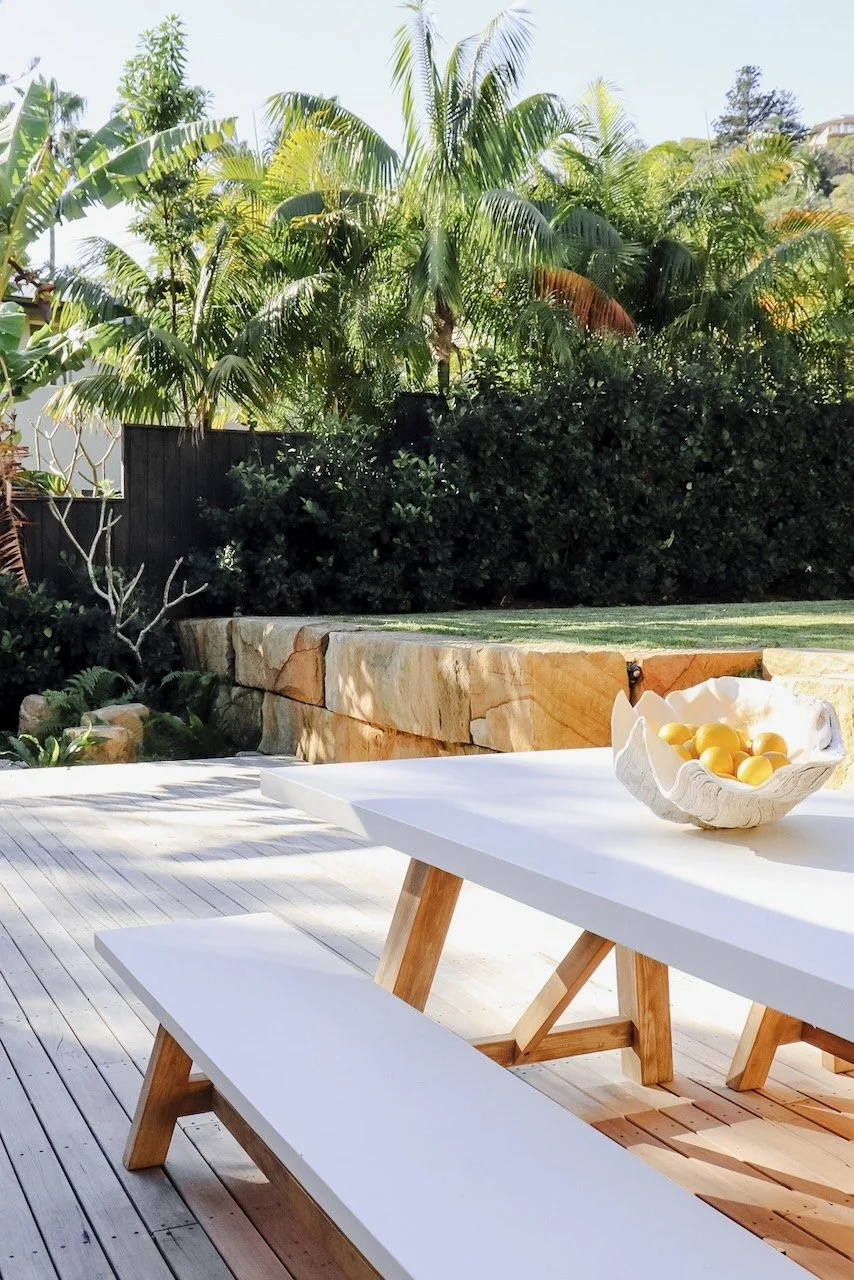 Outdoor patio with a white table, wooden bench, and a decorative bowl of lemons, surrounded by lush green tropical plants and trees.