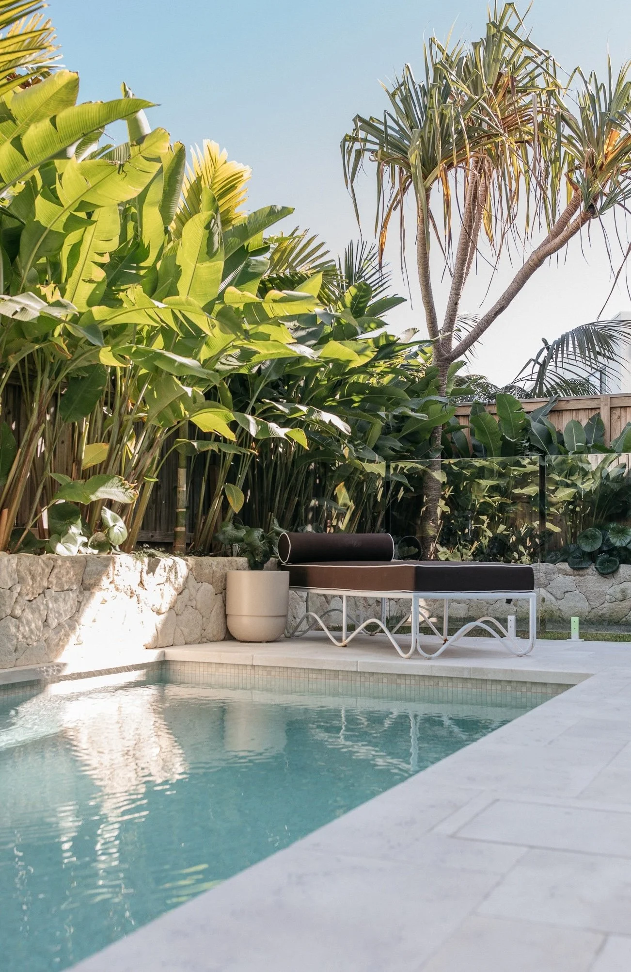 View of a backyard with a swimming pool, surrounded by lush tropical plants and trees, with outdoor furniture placed along the poolside.