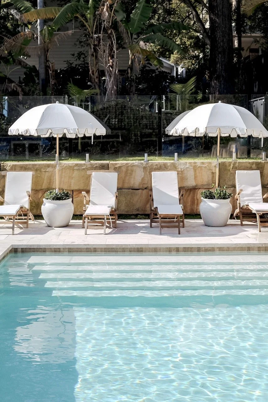 A backyard pool area with four white lounge chairs and two white umbrellas, with potted plants and a stone wall in the background.