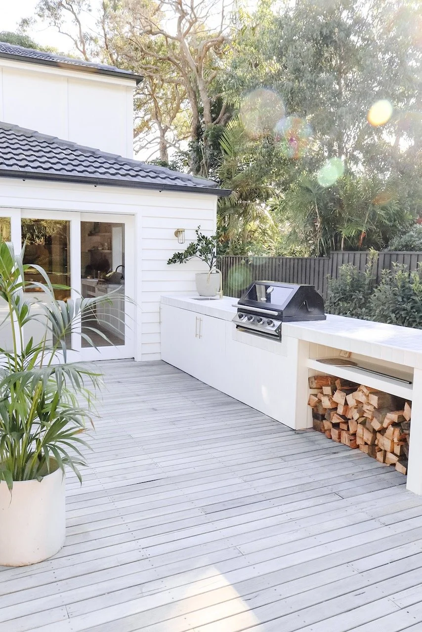 Outdoor patio area with a built-in grill, stacked firewood, a potted plant, and a white house with sliding glass doors surrounded by trees and greenery, illuminated by sunlight.