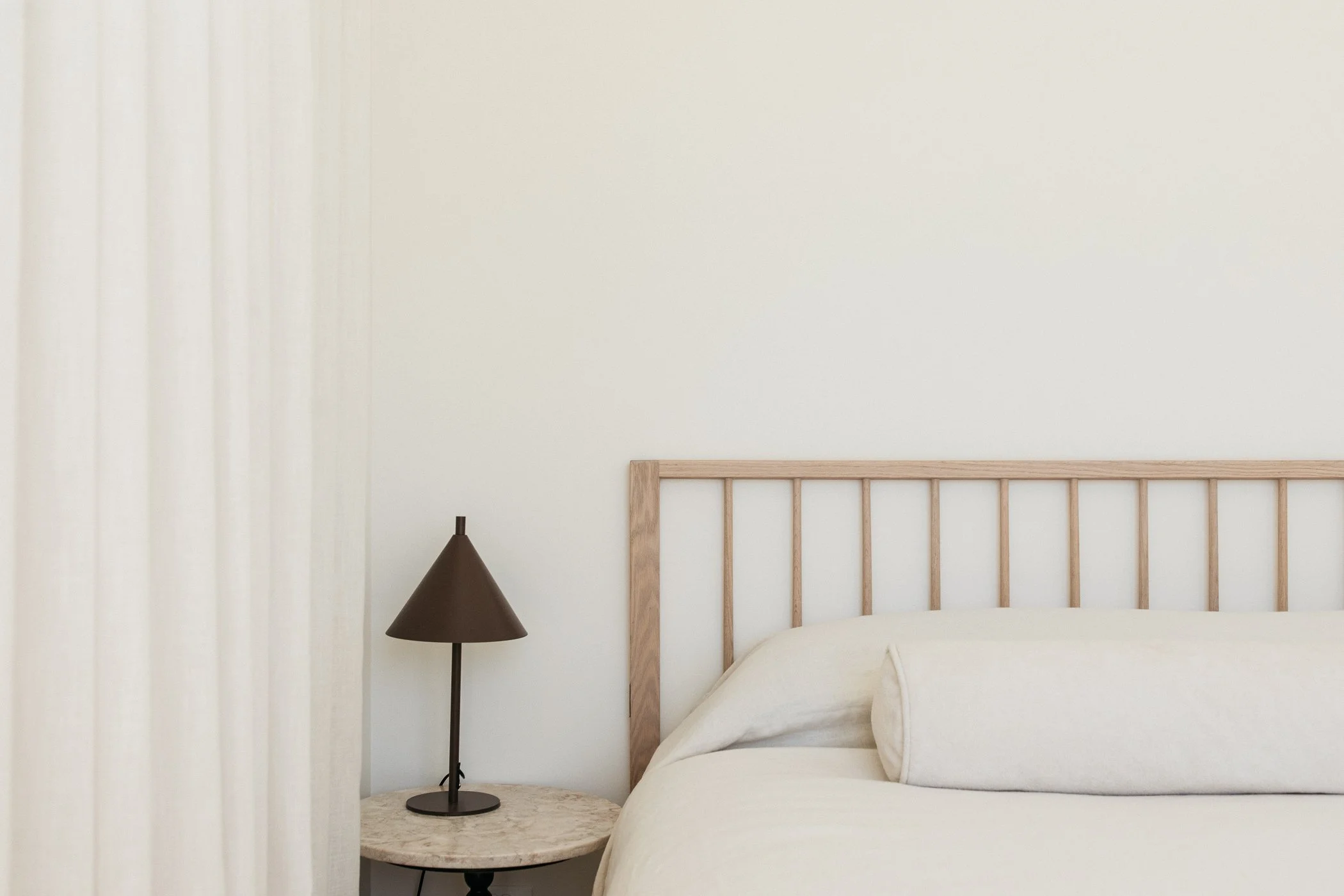 A bedroom scene featuring a wooden bed frame, a white bed with pillows, a black table lamp on a marble bedside table, and cream-colored curtains against a cream wall. Minimalist design.