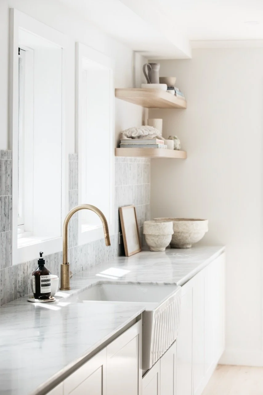 A minimalist kitchen with white cabinetry, marble countertops, and a single-handle brass faucet. Open wooden shelves hold decorative items and books, with a window casting natural light.