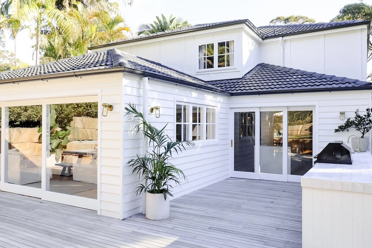 Backyard deck with white house, sliding glass doors, potted plants, outdoor grill, and surrounding trees.