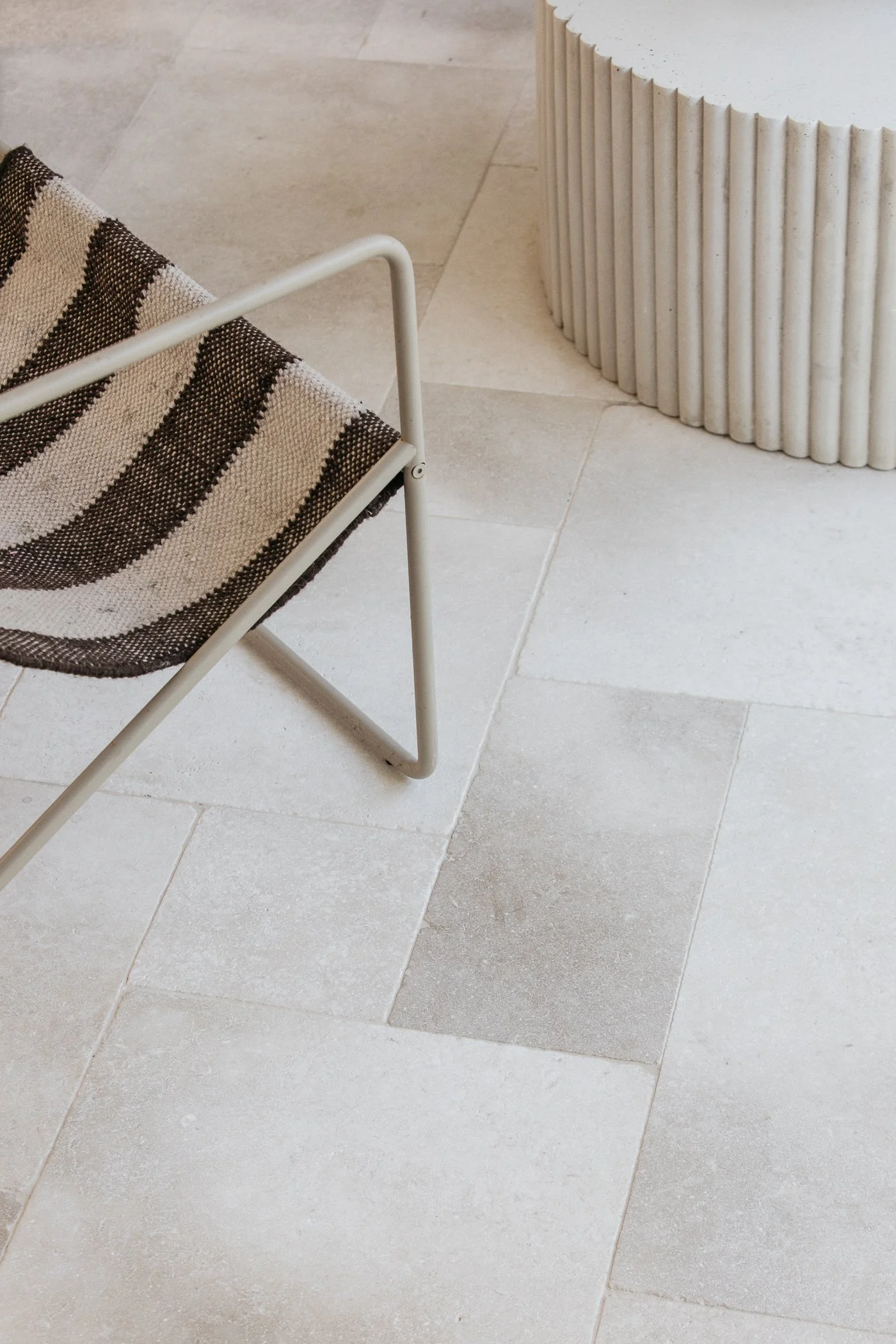 Close-up of a modern indoor space with beige tile flooring, a striped fabric lounge chair with a white metal frame, and a textured white cylindrical side table with vertical ridges.