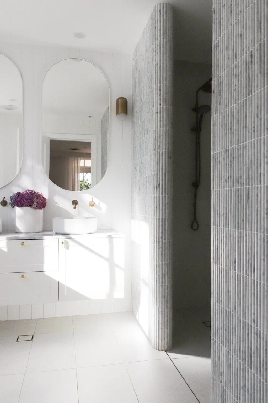 Modern bathroom with two white oval mirrors, a vase of purple flowers, a white countertop with a ceramic vessel sink, a brass wall-mounted faucet, and a walk-in shower with textured gray tiles and a showerhead.