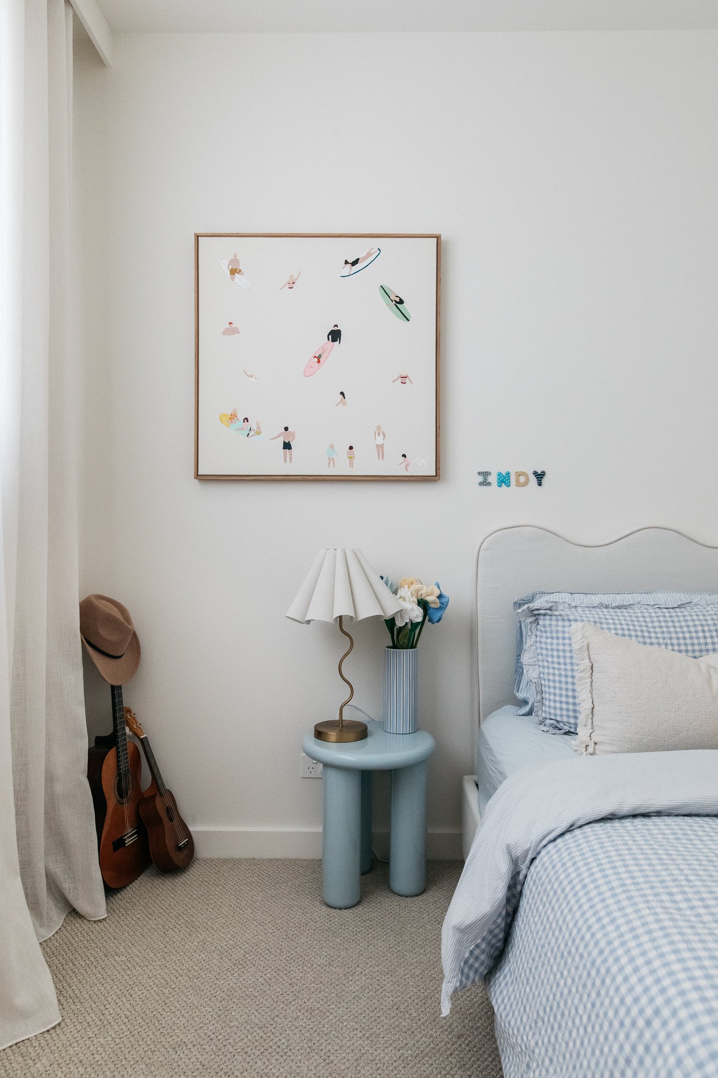 A bedroom with a bed, a blue side table, a white lamp, a guitar, and wall decor that spells out 'INDY'. The wall art features illustrations of people surfing.