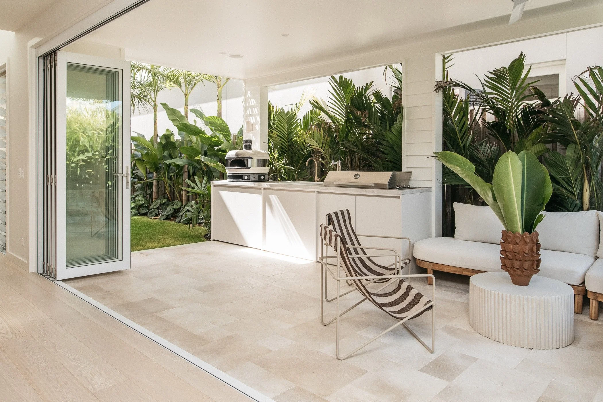 Modern indoor-outdoor living space with a sliding glass door, beige tiled floor, white furniture, a large potted plant, lush tropical greenery outside, and a built-in grill.
