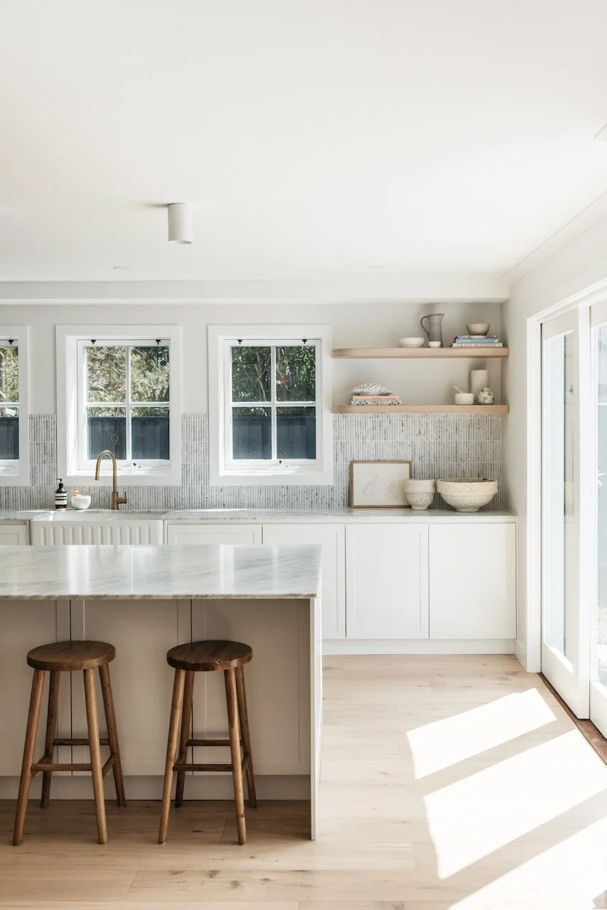 Bright, minimalist kitchen with white cabinets, a marble island, two wooden stools, three windows, and open wooden shelves with various bowls and dishes.