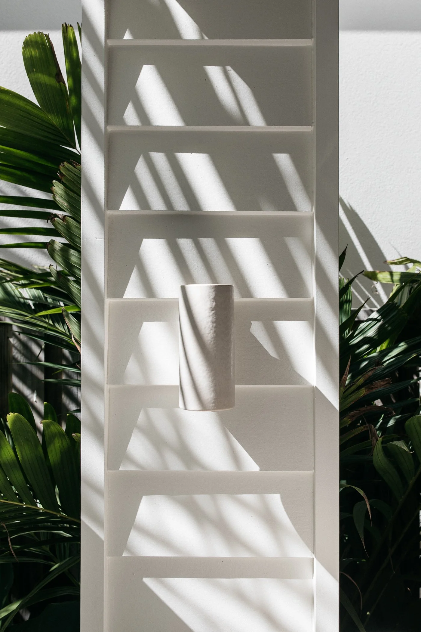White wooden shelf with horizontal slats, with a small white vase on the bottom shelf, surrounded by green tropical plants, illuminated by sunlight creating shadows on the wall.