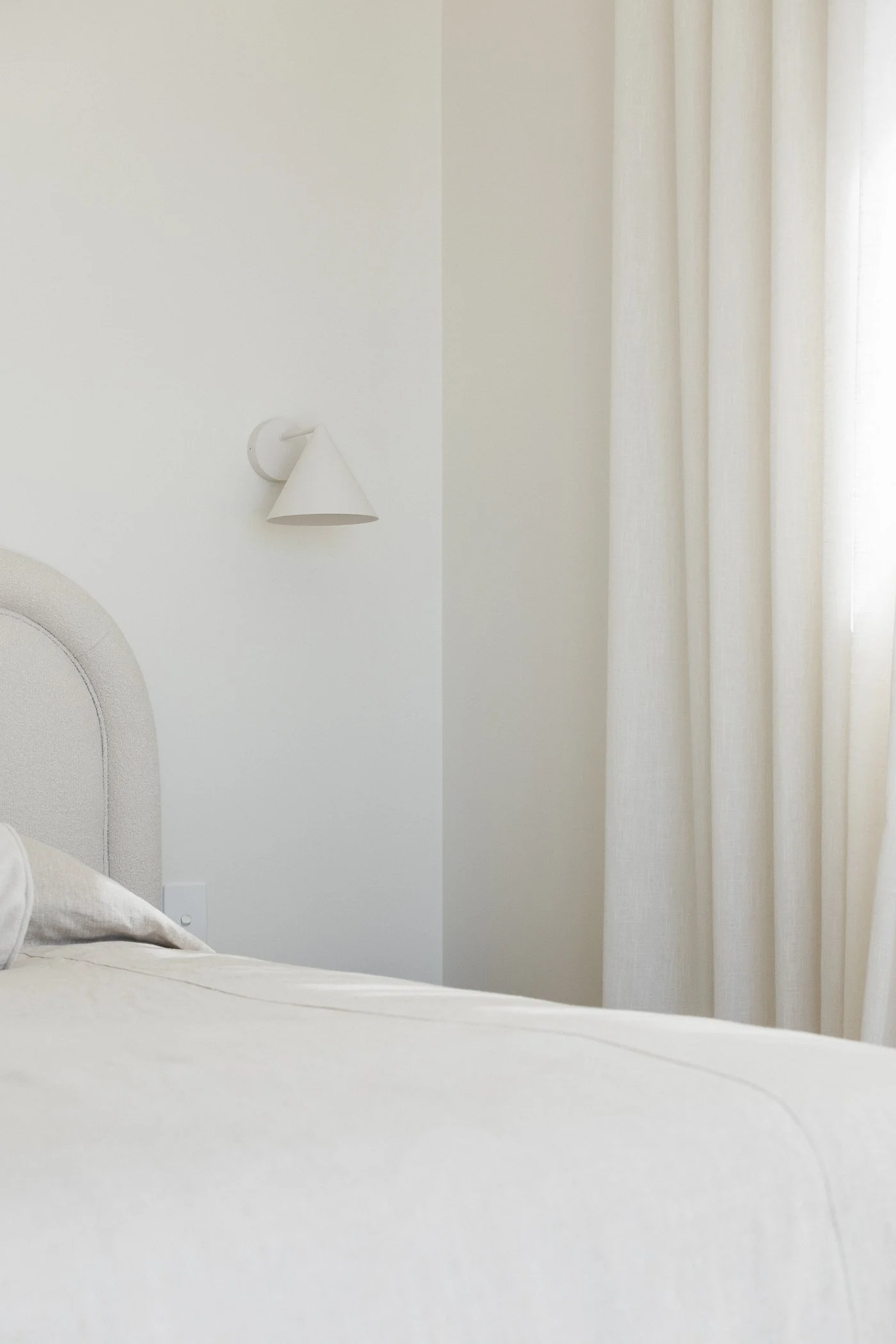 A bedroom with white walls, a white bed with a pillow, a wall-mounted white lamp, and white curtains on a window.