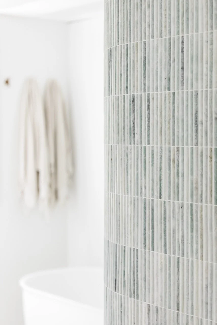 Close-up of a curved section of a wall covered with small rectangular marble tiles in shades of gray and white, with a bathroom and towels visible in the background.