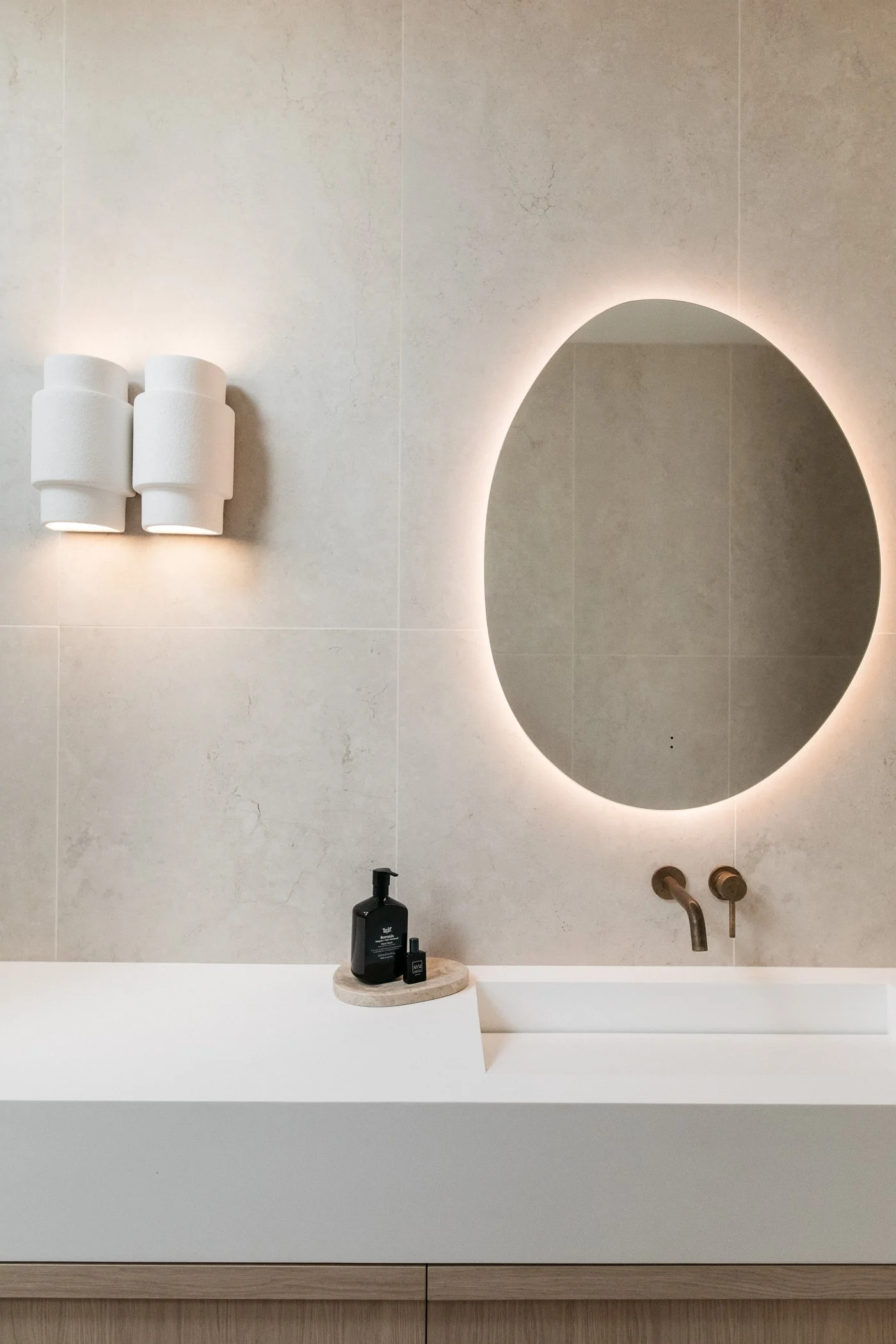 Modern bathroom with a backlit oval mirror, beige tiled wall, white countertop with black pump soap dispenser, and beige cabinetry below.