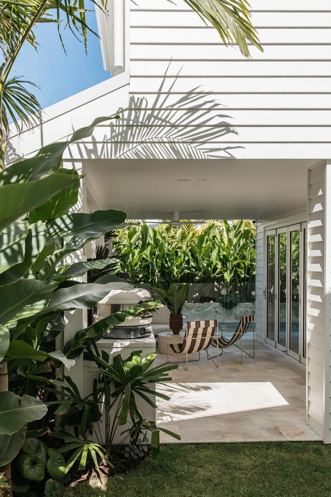 A patio area with lush green plants in the foreground, white siding on the house, and a small swimming pool with lounge chairs and a striped chair, shaded by a pergola with shadows of palm leaves on the wall.