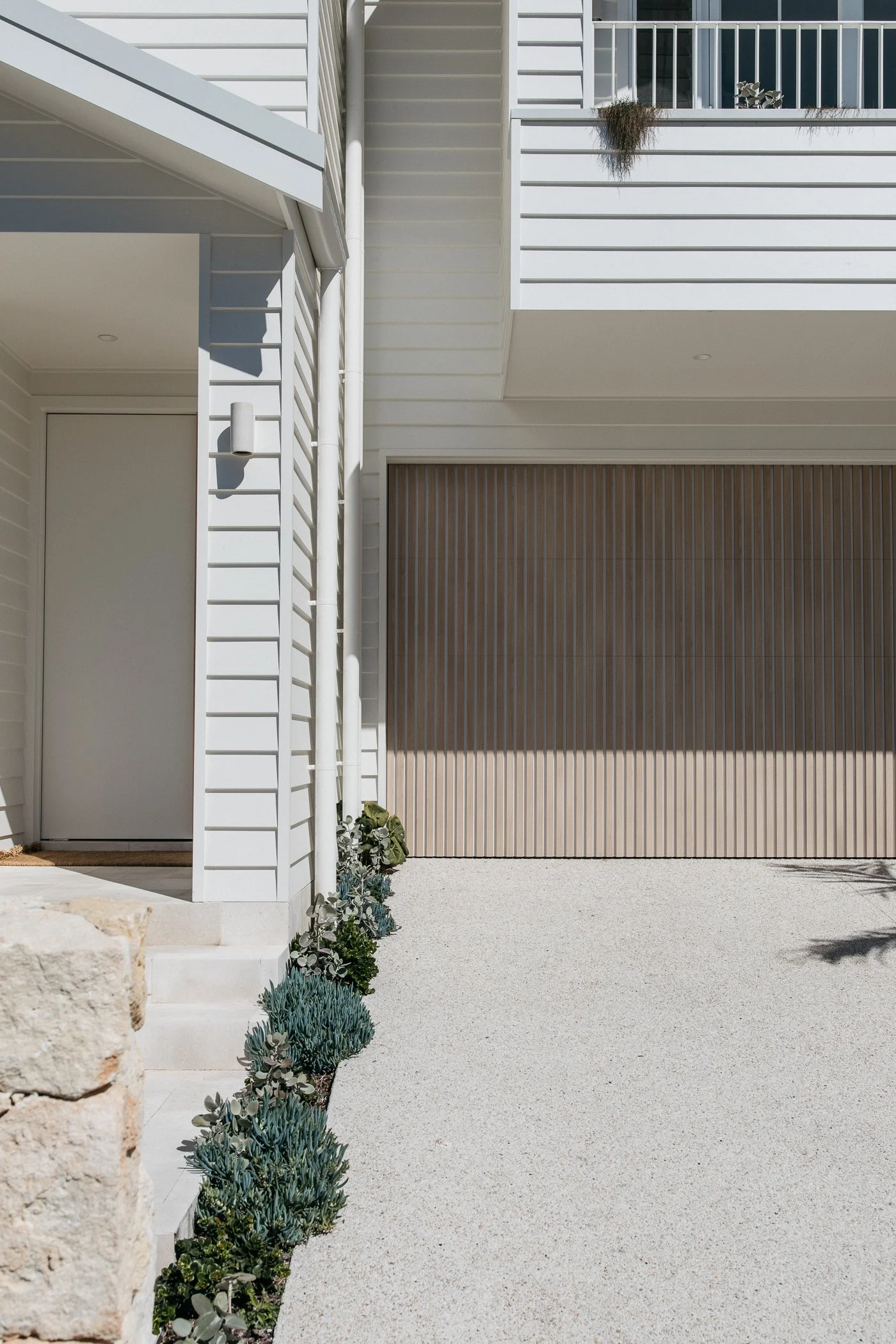 Close-up of a modern house exterior featuring white siding, a wooden garage door, a small planter with greenery, and a gravel driveway.