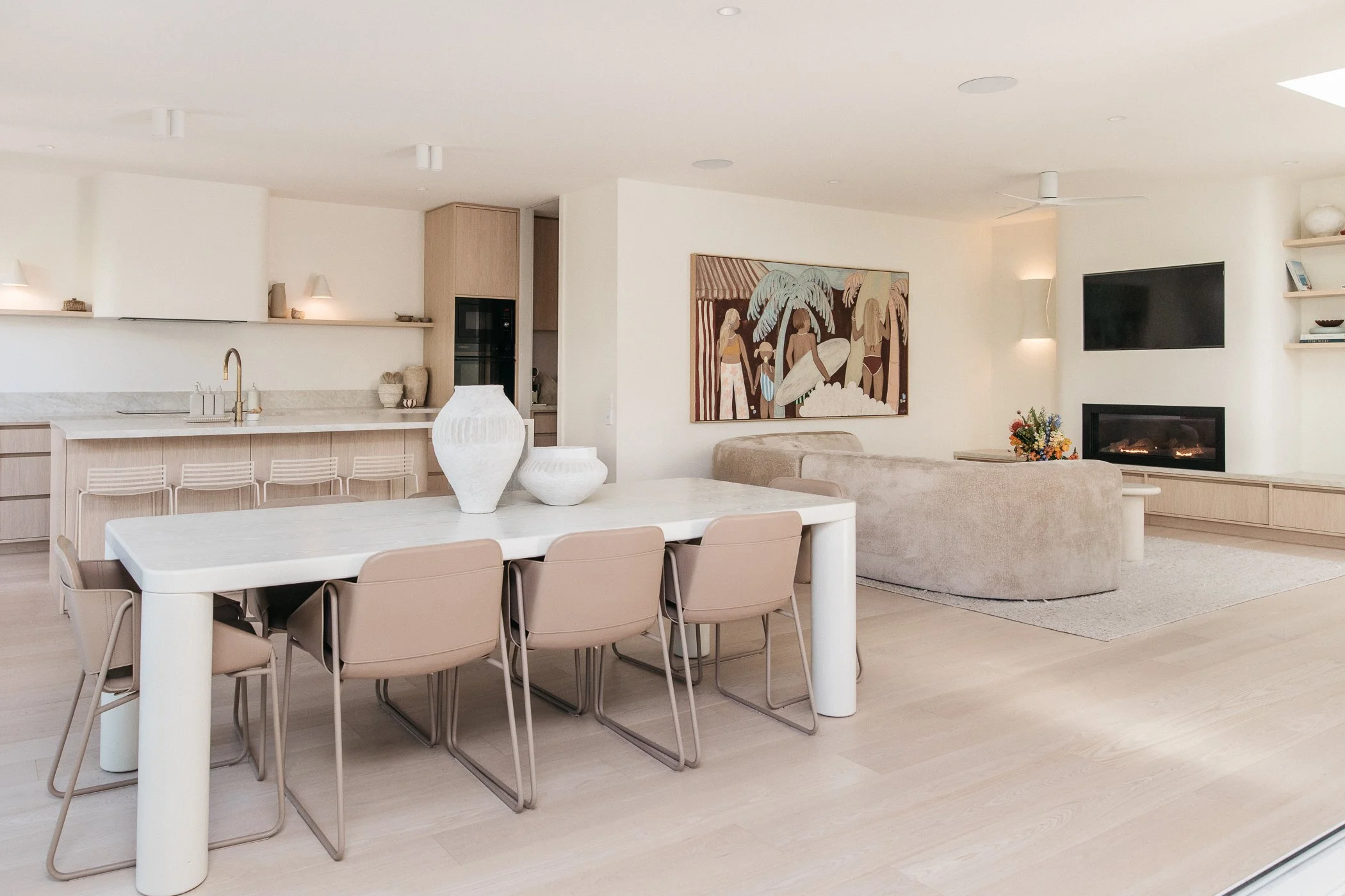 Modern, minimalist living room with white walls, beige furniture, a mounted flat-screen TV, abstract painting, and decorative items like vases and plants.