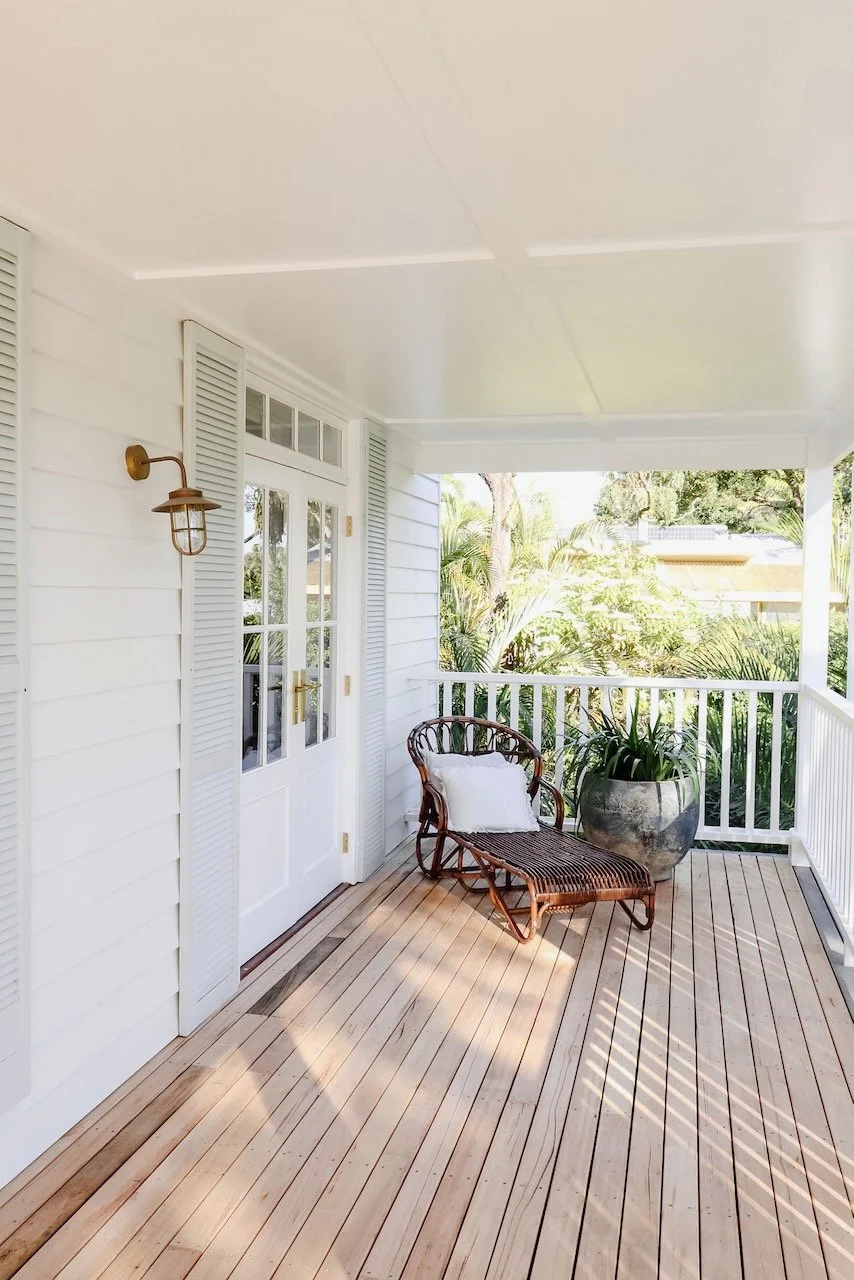 Front porch with a wooden chair, pillow, large potted plant, white siding, and door with glass panes.