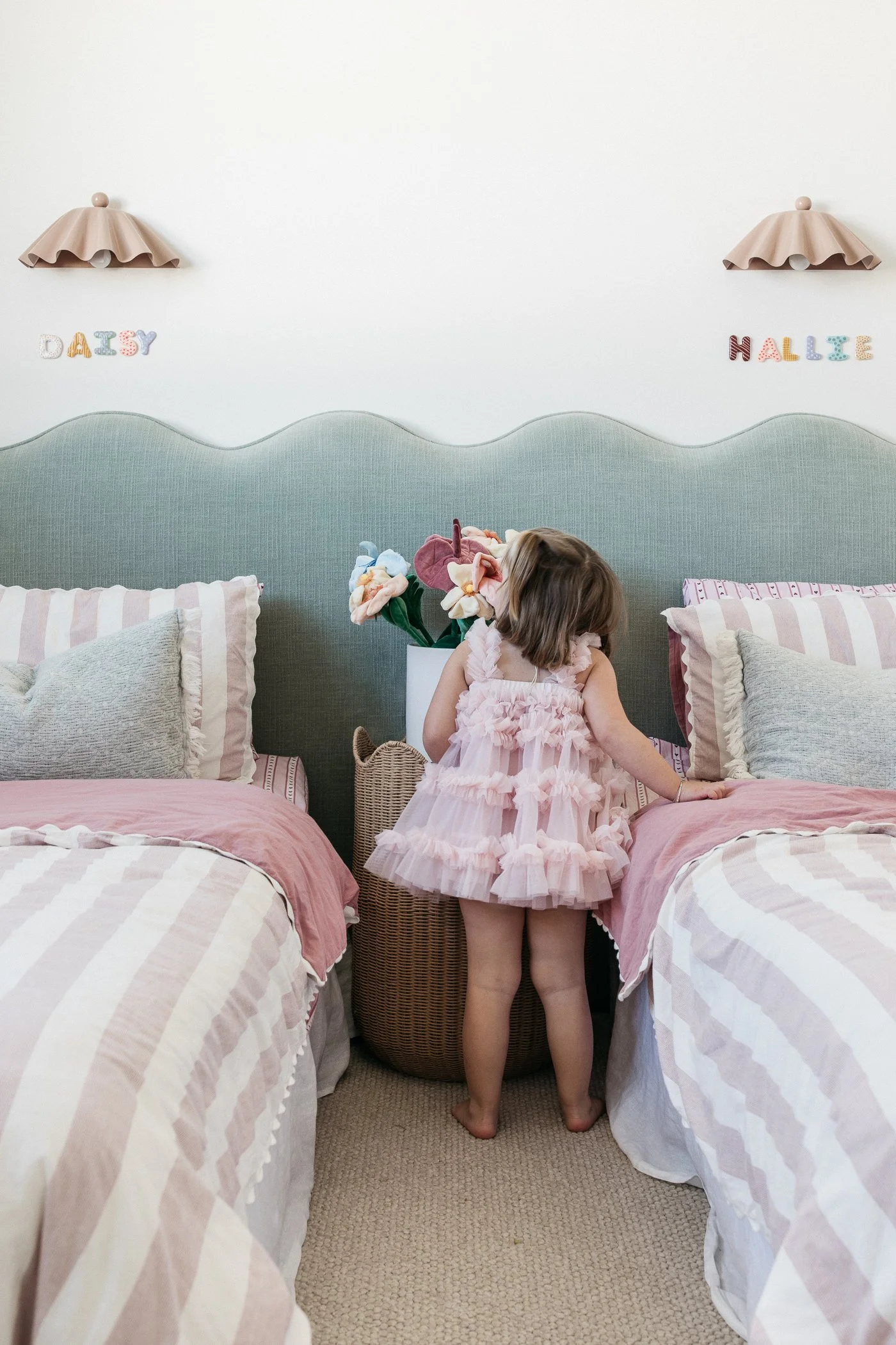 A young girl in a pink frilly dress stands between two beds with pink and white striped bedding, facing away from the camera. She is touching a wicker basket, and her head is turned to the side, with a partial view of her face and hair. The bedroom h