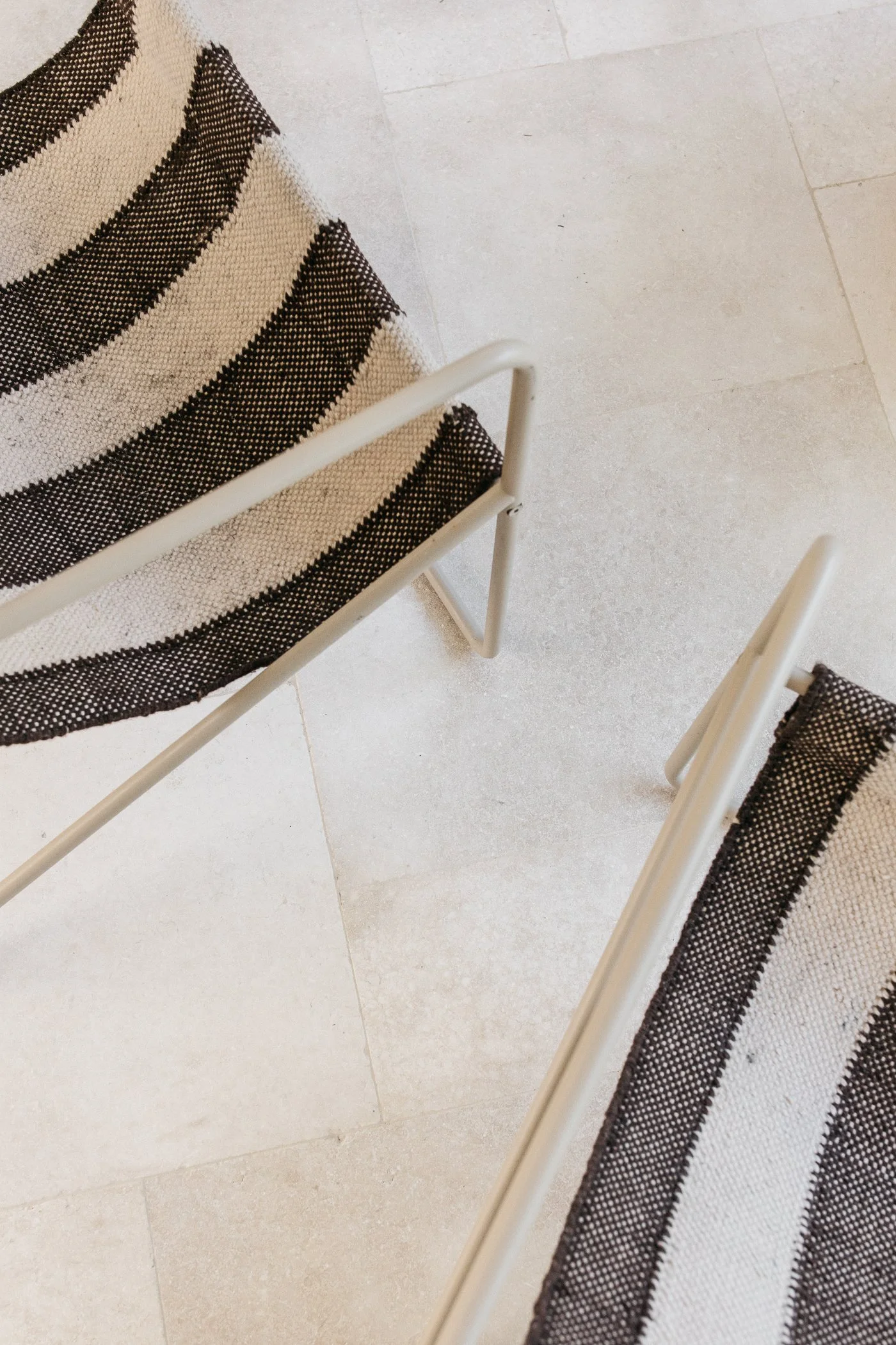 Close-up of two modern chairs with black and white striped fabric seats and white metal frames, placed on a beige tiled floor.
