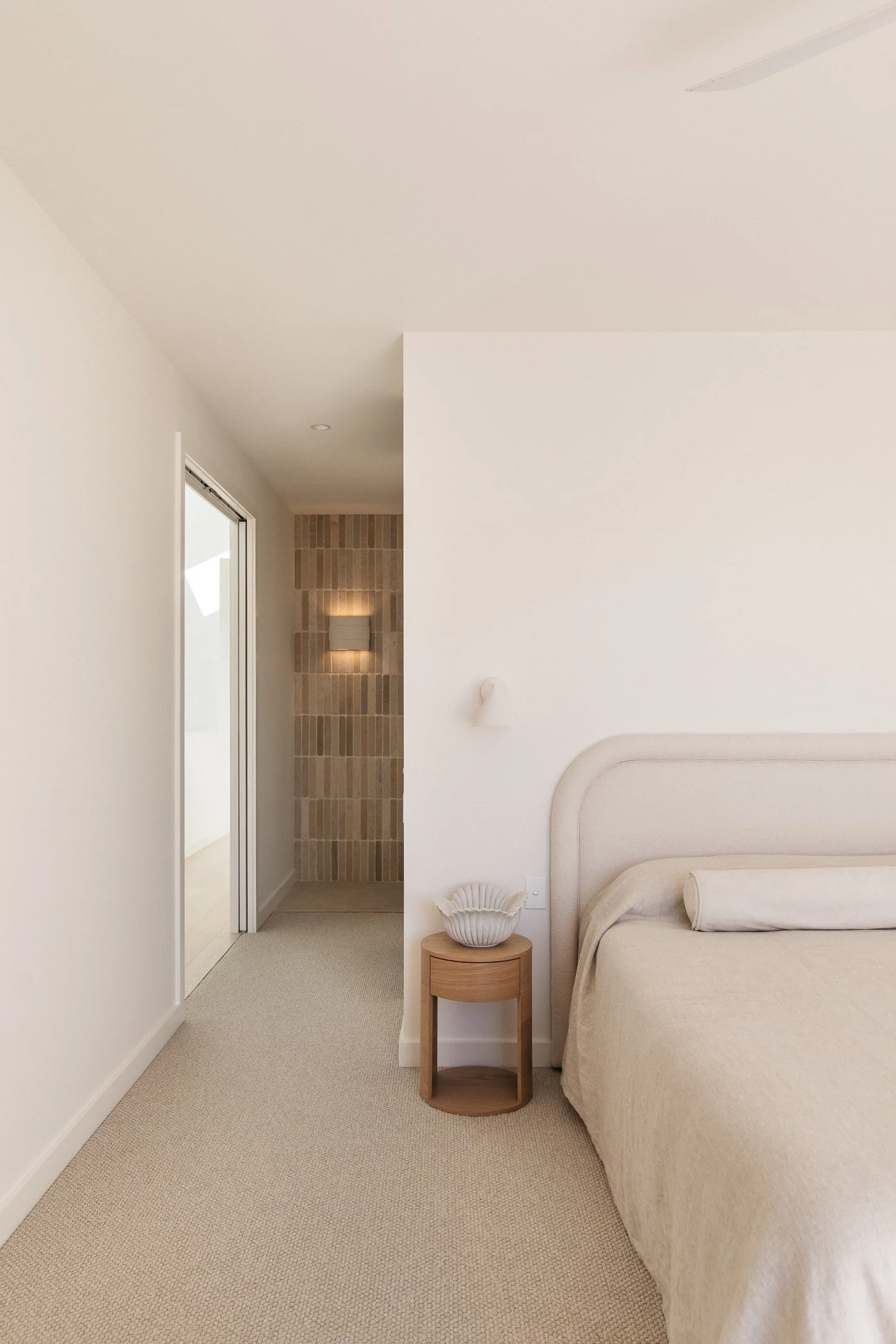 A minimalist bedroom with cream-colored walls and a beige bedspread. There is a small wooden side table with a decorative white bowl on it, and a curved headboard. The floor is carpeted in a light beige color. In the background, there is a doorway le