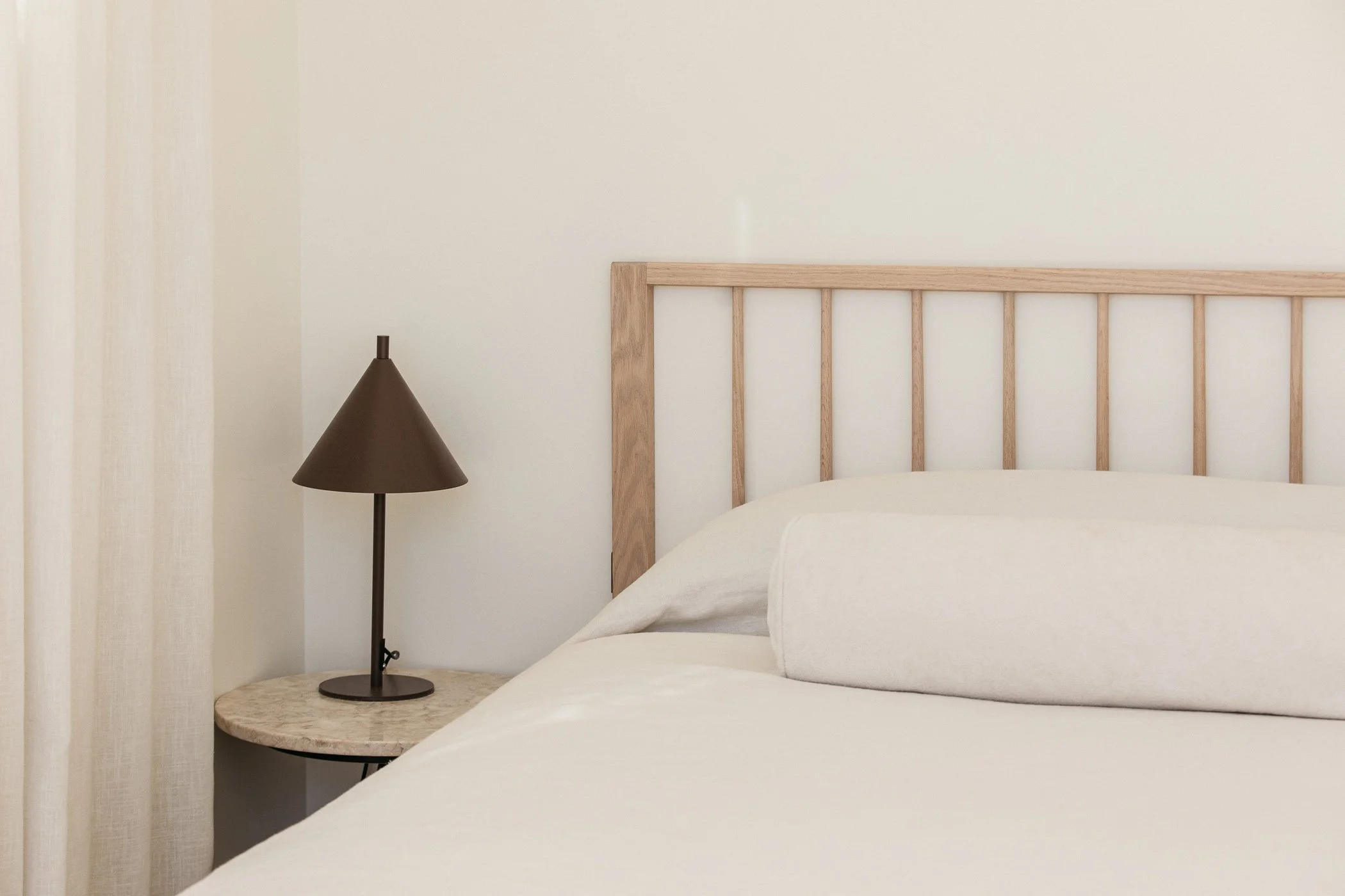 A bedroom with a wooden bed frame, a round marble side table, a brown table lamp, and a cream-colored bed with rolled pillows, minimal decor, and neutral tones.
