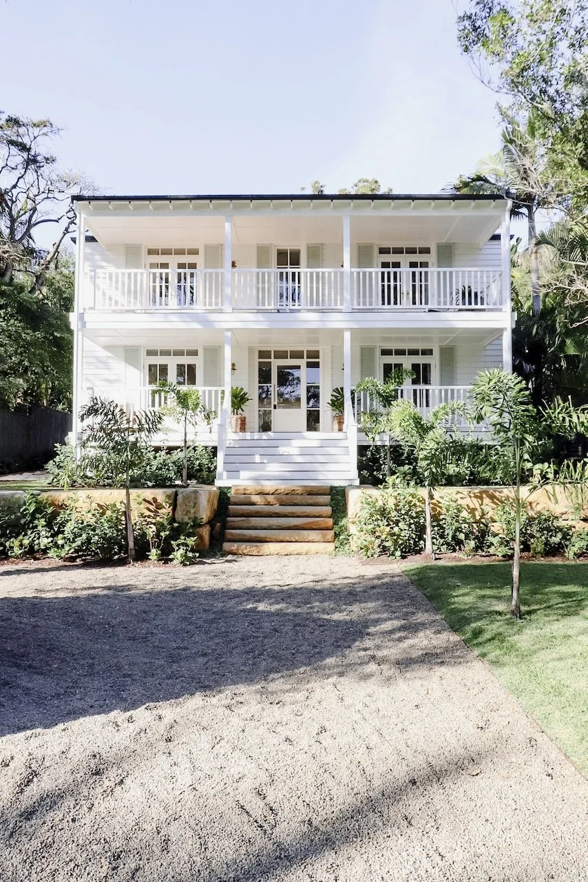 A two-story white house with a front porch and balcony, surrounded by trees and landscaped garden, with steps leading up to the entrance.
