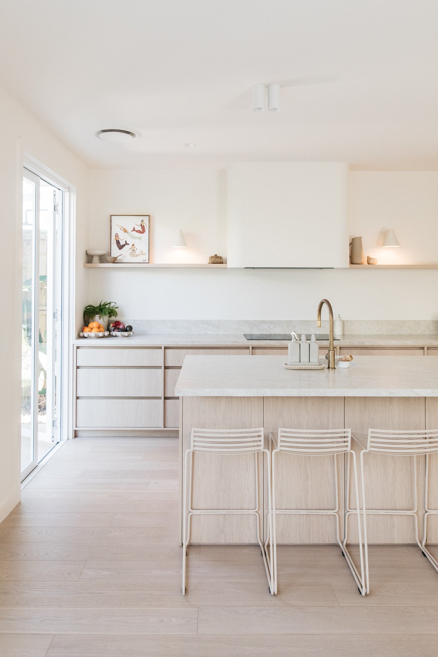 A modern and minimalist kitchen with light wood cabinets and flooring, white walls, a white island with a sink and gold faucet, white wire chairs, a fruit bowl with oranges and berries, a sliding glass door, artwork on the wall, and decorative items 