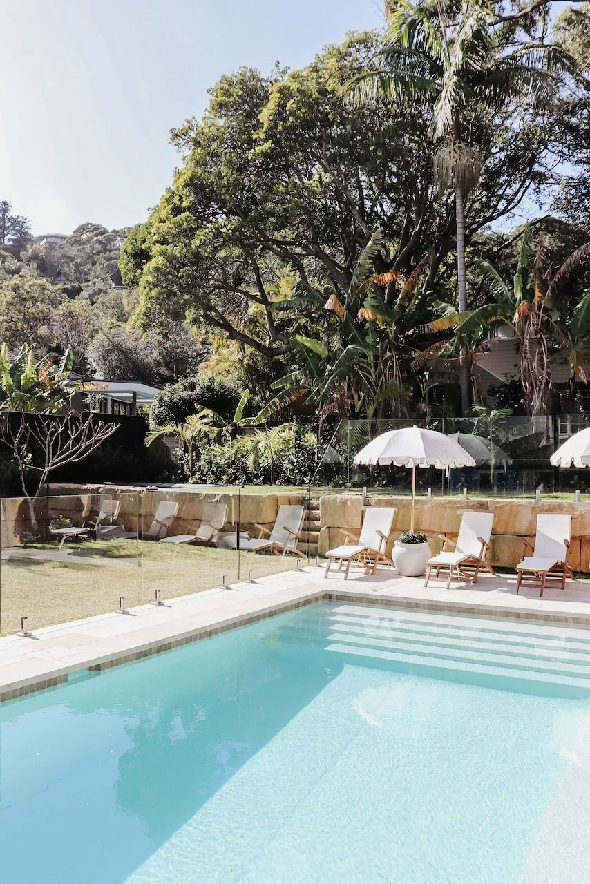 A backyard with a swimming pool, lounge chairs, and umbrellas surrounded by lush green trees and plants.