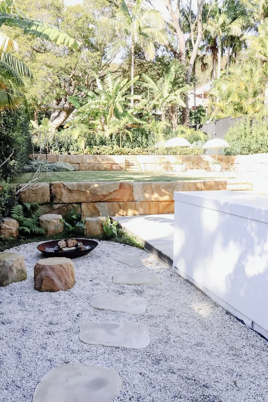 A backyard with a stone pathway on white gravel leading to a patio area with outdoor furniture and umbrellas, surrounded by lush green trees and plants.