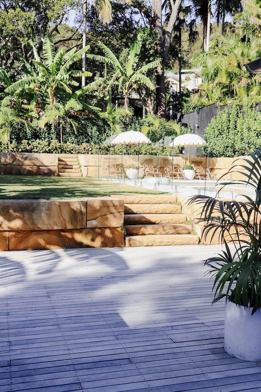 An outdoor backyard with wooden deck flooring, stone steps, lush green trees and plants, two white patio umbrellas, lounge chairs, and Potted plants during daytime.