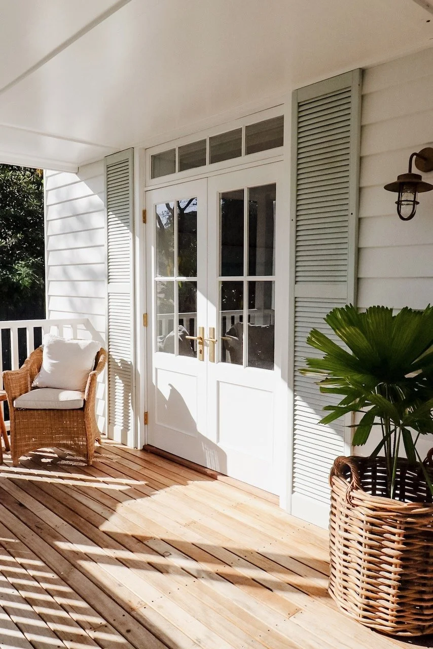 A porch with a white door, wicker chair with cushion, large potted plant, and wooden floor with shadows from the railing.