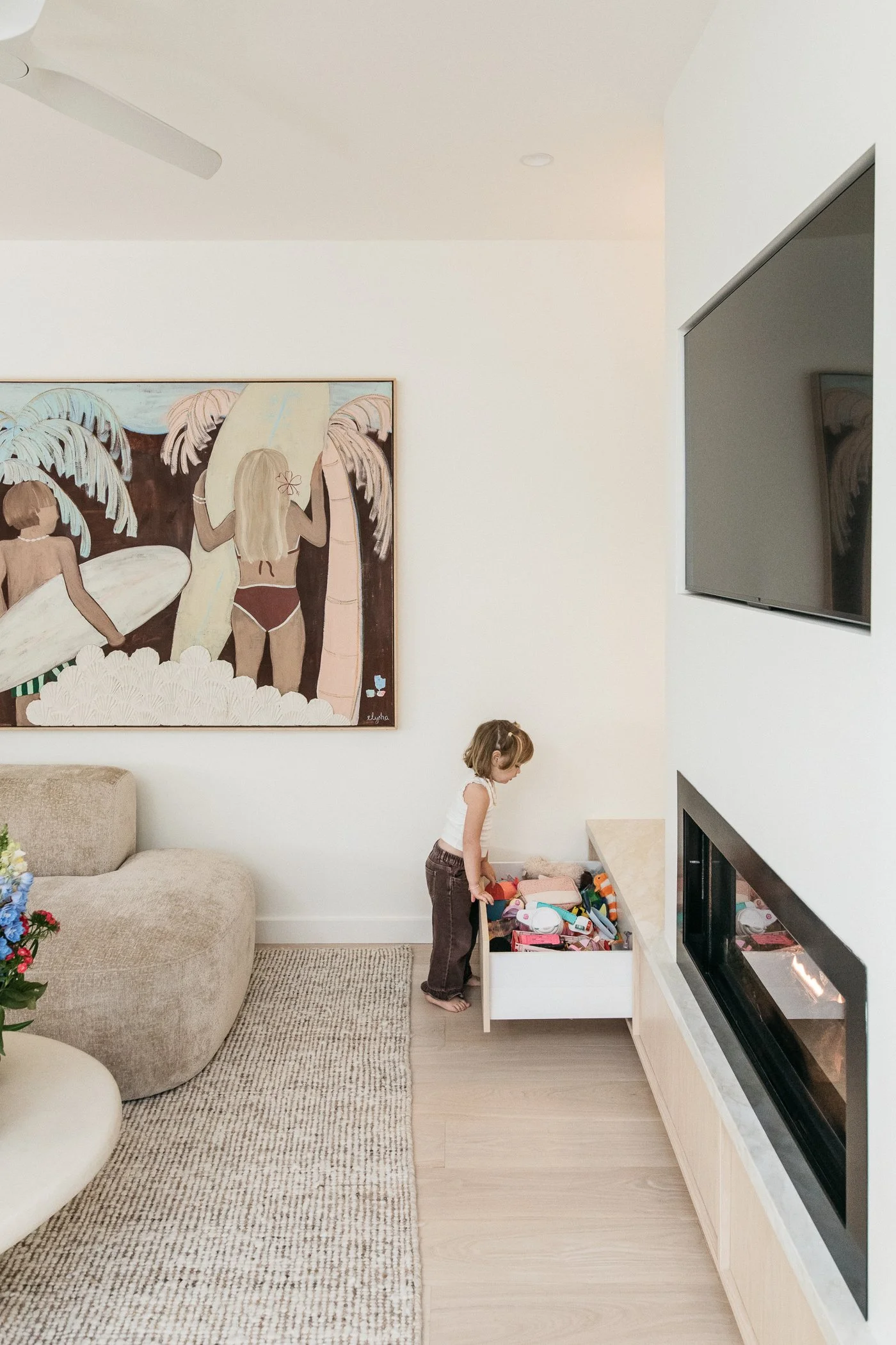A girl with short brown hair, wearing a white sleeveless top and brown pants, standing and looking into an open toy storage drawer filled with toys in a living room with a beige sofa, a rug, a large painting on the wall, a TV on the wall, and a firep
