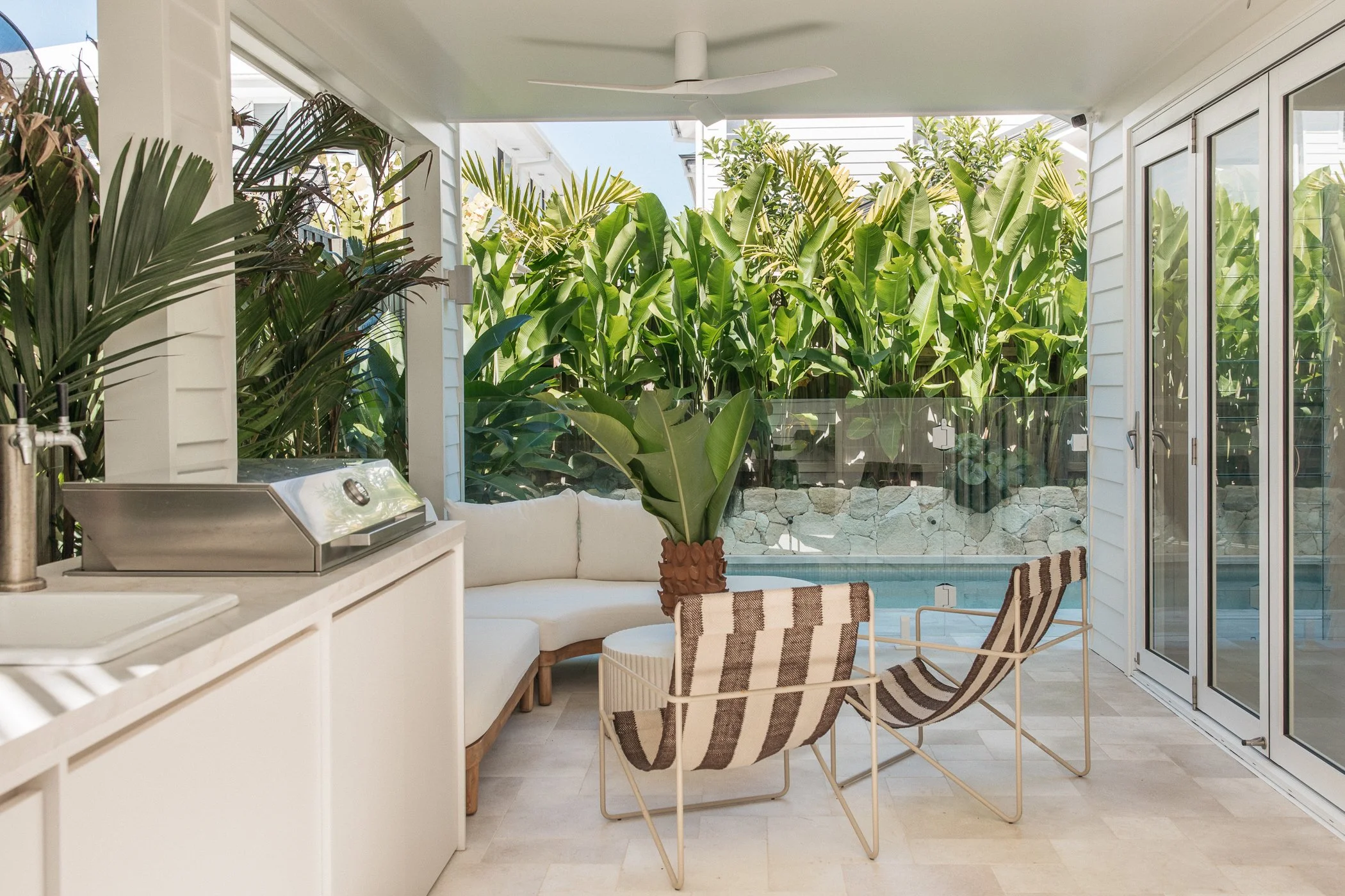 A covered outdoor patio with tropical plants, including banana and palm trees, and glass doors leading outside. There is a built-in grill or outdoor kitchen area, and seating with striped cushions, a small round table, and a ceiling fan.