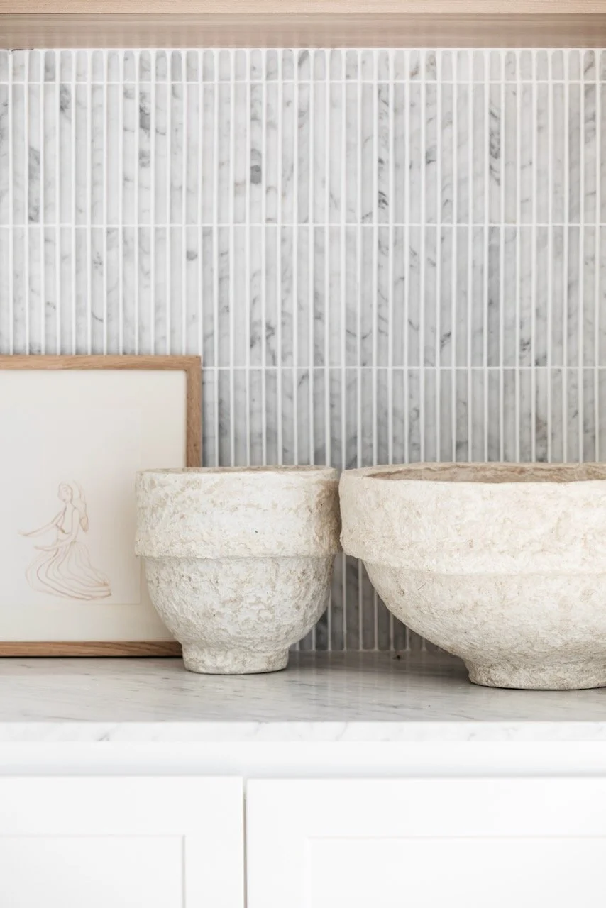 Decorative display of two textured white stone bowls of different sizes, a framed minimal drawing of a woman in a dress, and a marble countertop against a marble tile wall with white vertical bars.